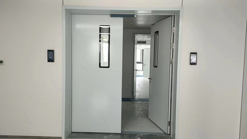 Light grey automatic sliding medical door with a tall narrow vertical glass observation panel sits in a pale metal frame beside an open matching hinged door. Black access control readers mount on plain off-white walls, looking through to an empty bright clinical corridor.