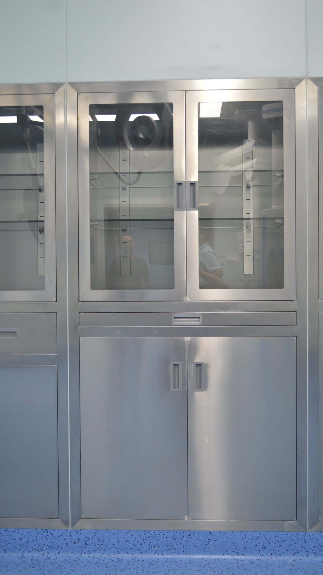Une armoire médicale en acier inoxydable brossé se dresse contre un mur gris-bleu pâle, au-dessus d'un sol bleu moucheté. Les portes vitrées supérieures, dotées de poignées encastrées, laissent apparaître des étagères intérieures en verre ainsi que les reflets de luminaires fluorescents, d'équipements et de personnes. Un tiroir sépare la partie vitrée de deux portes inférieures pleines en acier aux poignées assorties. Les surfaces stériles et réfléchissantes évoquent un environnement clinique propre.