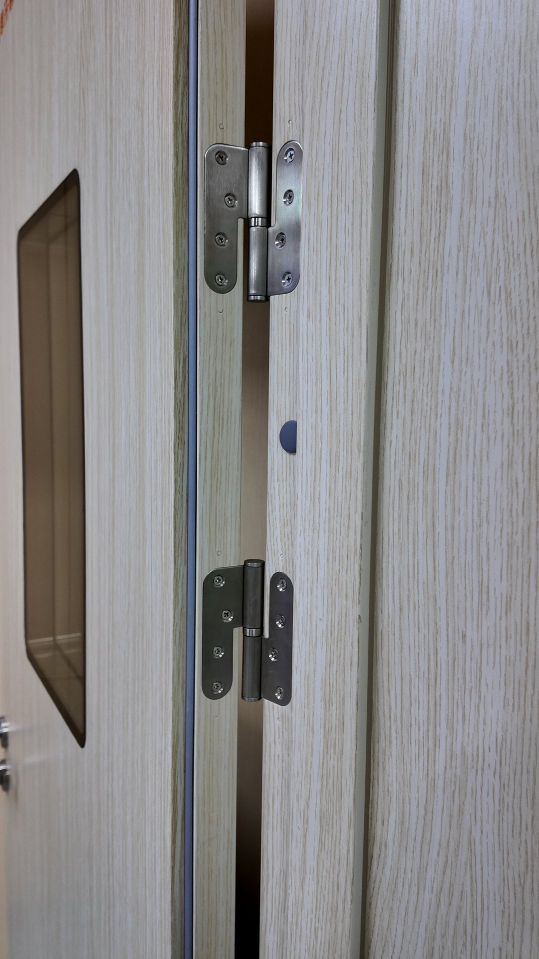 Close-up vertical shot of the hinged edge of a slightly ajar interior door, faced in pale cream laminate printed with fine straight pale oak woodgrain. Two matching brushed stainless steel hinges are spaced vertically in the narrow gap between door and matching woodgrain frame, each leaf secured with four Phillips head screws. A thin dark blue rubber seal runs along the frame edge, with a small round grey bumper dot mounted on the door edge between the hinges. A tall smoked amber translucent viewing window with angled corners appears on the door panel at left, near a small section of silver lock hardware. Soft neutral indoor light highlights the brushed metal texture and subtle woodgrain, with dark shadowed space visible through the narrow door gap.