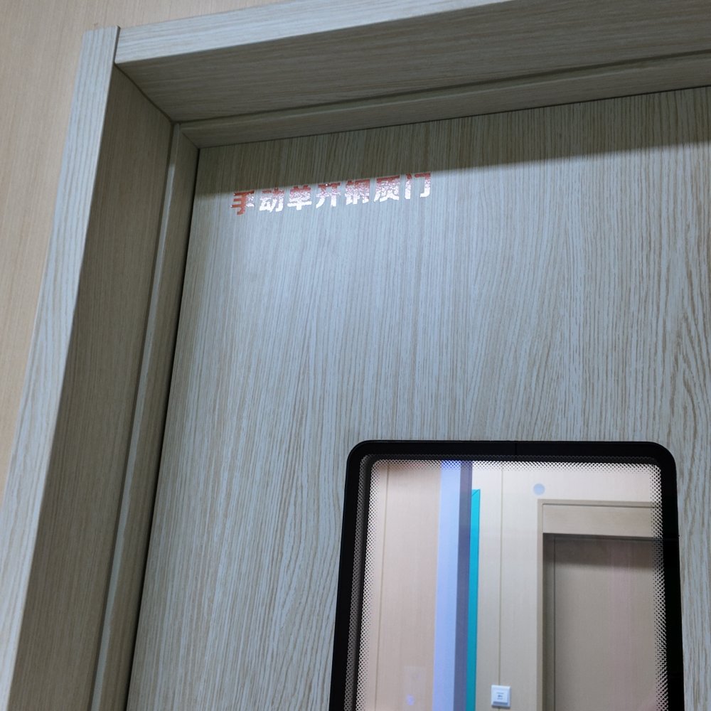Upward angled close-up of a pale wood-grain manual single-swing steel interior door, set in a matching wood-look frame surrounded by light wood finished wall panels. Shiny metallic red Chinese characters labeling the door type sit on its upper section. A narrow rounded rectangular observation window with a glossy black frame and dotted gradient border reveals a beige clinical corridor beyond, holding another matching door, silver pull handle, white light switch, small blue-lit control panel, and bright turquoise vertical wall stripe. Soft even diffused indoor light illuminates the smooth uniform surfaces.