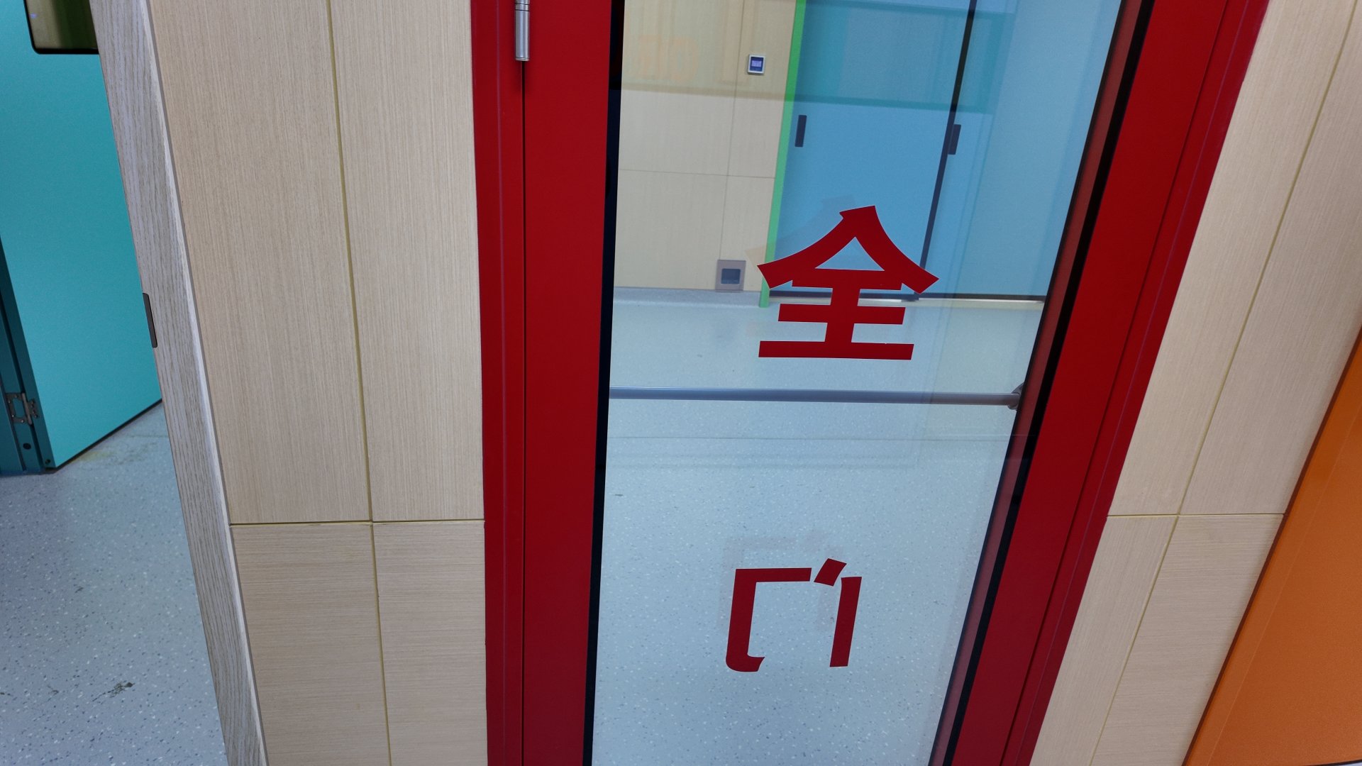 Clean modern indoor corridor with light wood-grain wall panels and pale grey speckled seamless flooring, lined with brightly painted doors. The prominent foreground door is a glossy red-framed full-length glass safety door, marked vertically with three bold red Chinese characters identifying it as a safety door, fitted with silver metal hinges along its left edge. To its right is a closed glossy bright orange double door with a keyed lock and hanging key, while through an open left doorway, a closed bright green sliding double door sits at the end of the adjacent hall beside a partially open turquoise door. Even neutral overhead lighting casts soft reflections on smooth door surfaces, and a small white access control panel is mounted beside the distant green door.