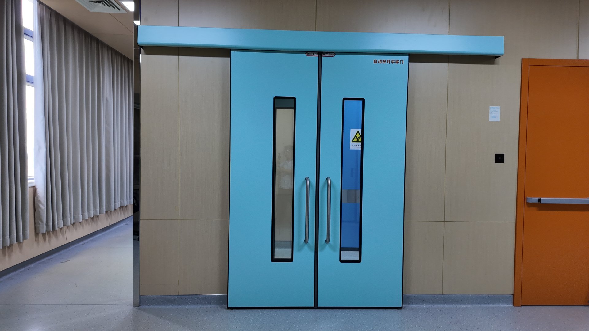 Light turquoise automatic double sliding radiation-shielded doors stand centered in a clean modern hospital corridor, set into beige wood-effect wall panels. Each door has a tall narrow black-framed vertical glass window, left clear and right tinted blue, with two long brushed stainless steel pull handles between them; the right door bears a yellow radiation trefoil warning sign, red Chinese text labeling the automatic sliding door, and AUTO direction markers at the top seam. A matching turquoise track header runs above, a bright orange fire door with silver panic bar sits to the right, and the corridor extends left past floor-length pale grey curtains, lit by even cool clinical light over smooth speckled grey flooring.