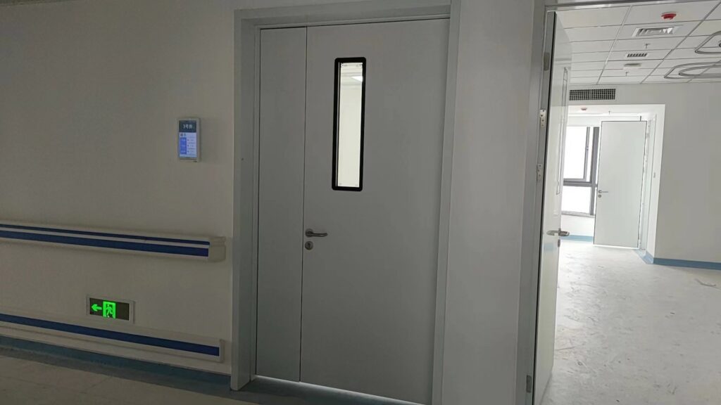 A sterile, newly finished hospital corridor with plain off-white walls and blue protective trim features a closed light-gray medical door at center, fitted with a tall narrow black-framed vertical window, a silver lever handle, and a round lock below it. 