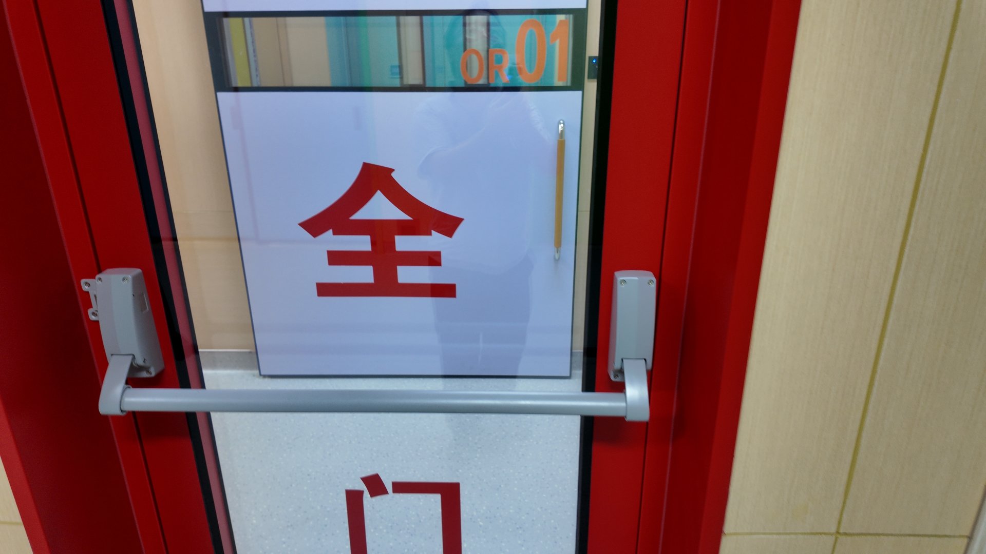 Straight-on close-up of a glossy bright red-framed glass hospital exit door, fitted with a horizontal matte silver panic push bar across its lower section. A frosted white opaque panel on the glass displays one large bold red Chinese character, with the top of a second red character cut off at the bottom frame edge. Above the panel, clear glass is marked with bright orange sans-serif text reading OR 01 for operating room 01. A slim light wood vertical handle with silver end caps sits behind the glass to the right, faint reflections of the photographer and muted teal and beige interior walls appear on the smooth glass surface, and pale beige wood-look tile wall cladding sits beside the frame under even, bright neutral indoor lighting.