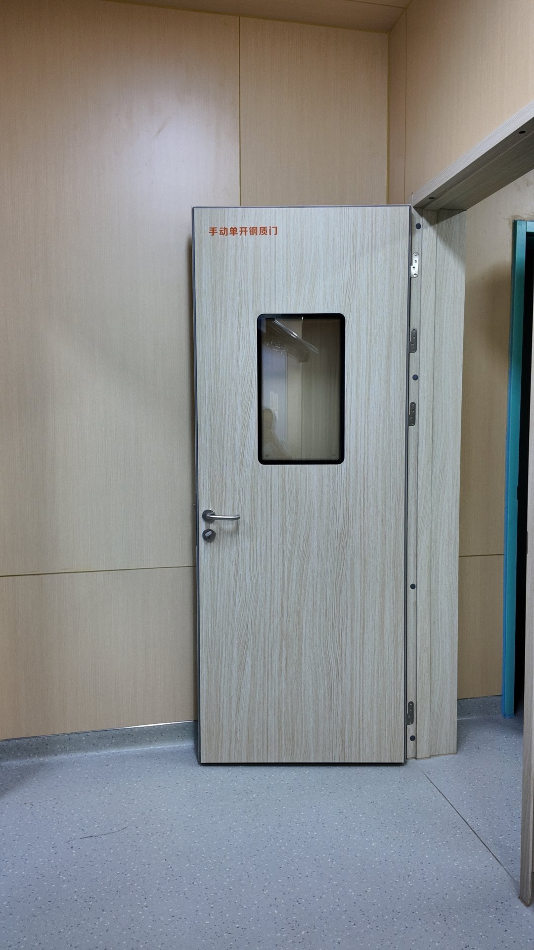 A closed light woodgrain manual single-swing steel door stands centered in a clean clinical hospital corridor corner. It has a small black-framed semi-transparent glass observation window at its midpoint, a brushed silver lever handle and key lock on its left, and exposed metal hinges along its right edge mounted to a matching woodgrain frame. Bold red Chinese text labeling the door type is printed at its upper left. Walls are clad in matching light beige wood paneling, the floor is smooth grey speckled non-slip commercial flooring, and a teal-trimmed adjacent doorway is visible at far right under even neutral indoor lighting.