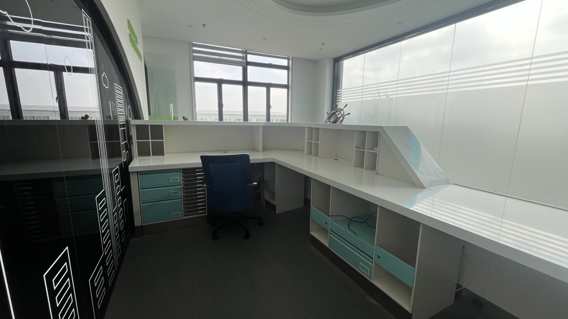 Bureau moderne vide et lumineux centré sur un comptoir d'accueil blanc brillant en forme de L avec des casiers ouverts, des fentes à dossiers et des tiroirs bleu sarcelle doux dotés de poignées argentées. Une chaise roulante bleu foncé est placée devant le bureau sur une moquette anthracite foncé. Une cloison incurvée en verre noir sur la gauche est ornée d'un dessin au trait blanc représentant des gratte-ciel, des fenêtres lointaines laissent entrer une lumière du jour diffuse et voilée, et un mur d'intimité dépoli à rayures longe le côté droit, avec un décor de barre de navire en métal et des murs blancs qui parachèvent cet espace minimaliste et épuré.