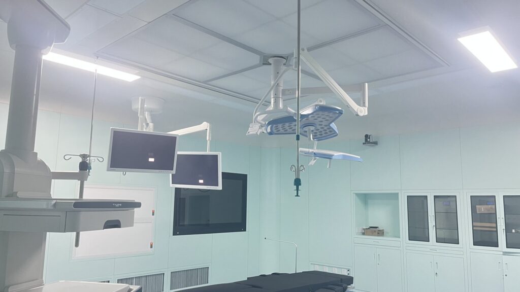 A clean, unused sterile modern operating room viewed beside the black padded operating table at the bottom of the frame, with pale mint-green seamless hygienic wall panels, a light grey segmented suspended ceiling, and bright flush rectangular LED panels providing even cool white illumination. Articulated white ceiling-mounted support arms hold two widescreen medical monitors with dark reflective screens, a large multi-bulb shadowless surgical light with pale blue trim, and IV poles with hanging hooks. The right wall features integrated pale cabinetry with dark glass doors, closed lower storage, an open recessed shelf holding a small cardboard box, and a small mounted security camera. A dark recessed display sits on the far wall, louvered ventilation grilles run along the wall base, and a tall grey equipment boom stands at the far left, with all smooth non-porous surfaces designed for easy clinical sterilization.