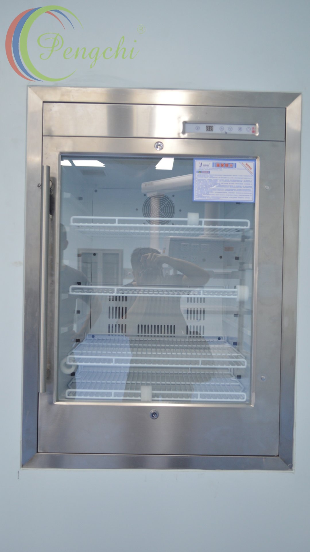 A recessed medical warming /cold cabinet framed in brushed reflective stainless steel sits flush in a smooth matte pale gray wall. It has a clear glass door with a long vertical stainless steel handle on its left edge, and two keyed locks above and below the glass panel. A narrow digital control strip at the upper frame right displays an 8.8 temperature readout beside function buttons, with a blue-bordered white Chinese warning label fixed to the upper right glass. The empty light gray interior holds four adjustable white coated wire shelves and a circular rear circulation fan above vertical vents, while the reflective glass shows faint reflections of the photographer, fluorescent ceiling lights, and surrounding clinical room features.