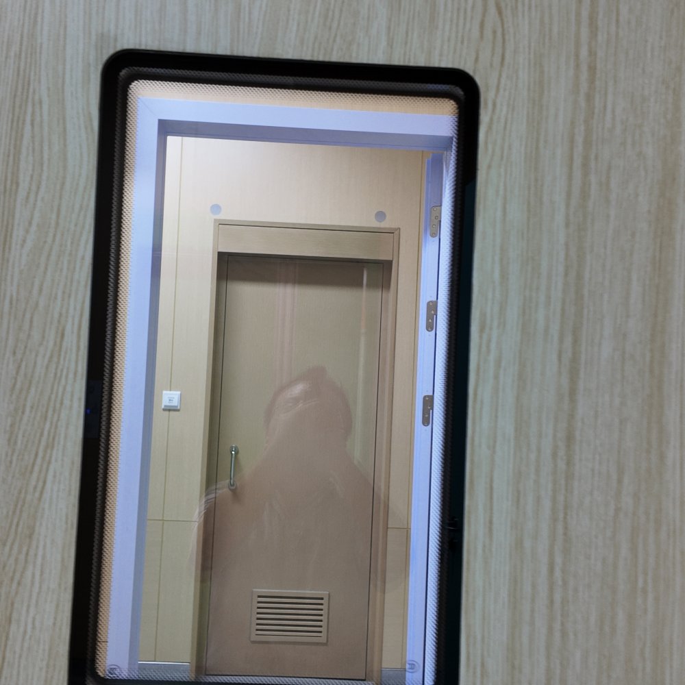 A rectangular rounded-corner window with a thick glossy black frame is set into a light beige vertical woodgrain-patterned wall. Through clear, slightly reflective glass, a neutral interior is visible, centered on a matching light wood door with a slim silver pull handle at left and a horizontal-slatted louver vent near its bottom. Three metal hinges line the door’s right edge, a small white light switch sits left of the door, and pale cool white trim outlines the doorway. The faint silhouette of the photographer is reflected on the glass, and the scene is lit by even, diffused neutral indoor light.