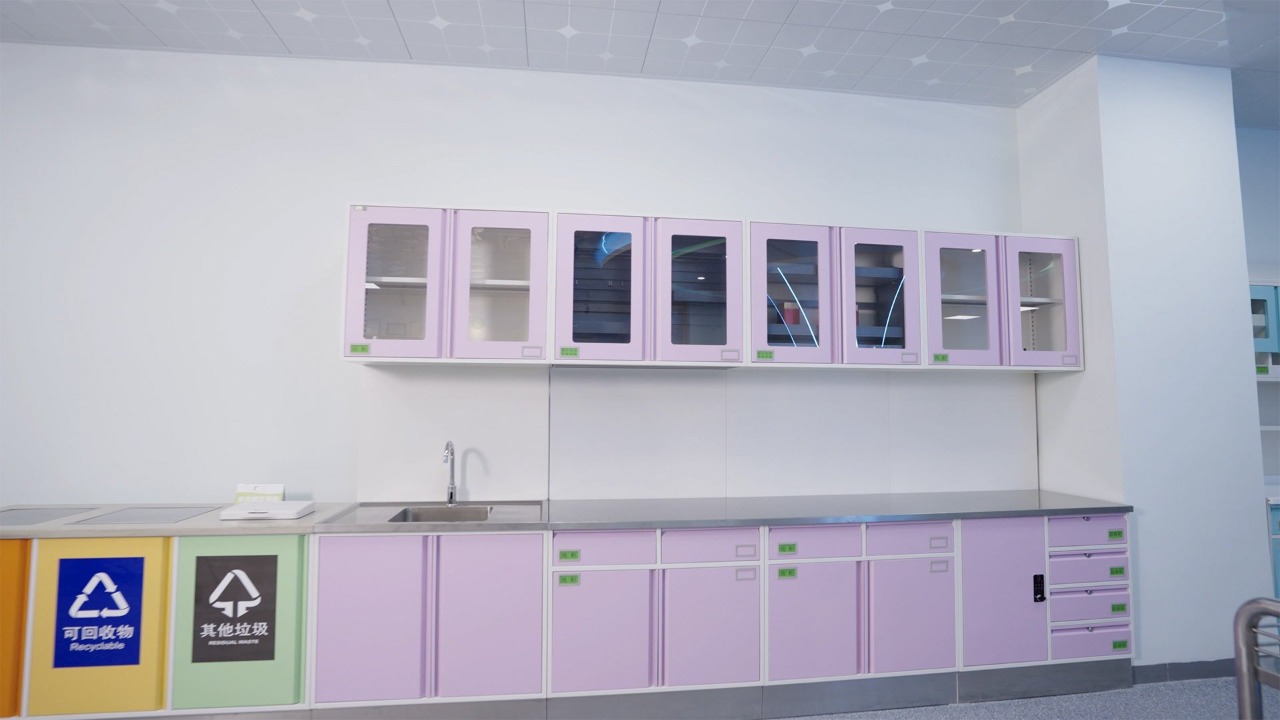 Gabinetes médicos de pared color lavanda con puertas de vidrio sobre un mostrador de acero inoxidable con fregadero y contenedores de reciclaje codificados por colores en una sala de clínica