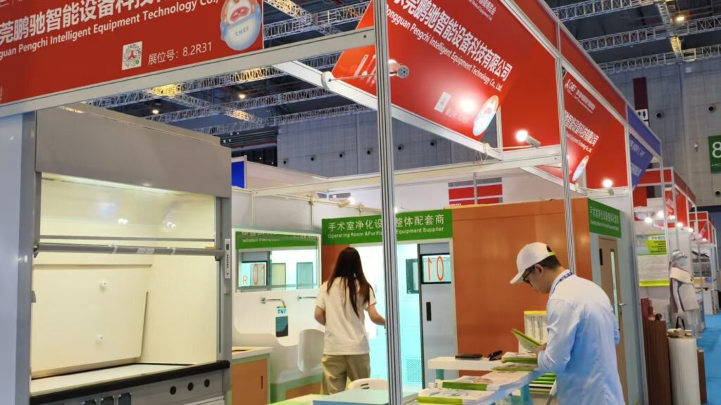 Una señalización superior de color rojo intenso indica el stand 8.2R31 de Dongguan Pengchi Intelligent Equipment Technology en una feria comercial médica en un centro de convenciones con cerchas metálicas vistas en el techo y paredes grises paneladas. Entre los productos expuestos se incluyen campanas de extracción para laboratorio de acero inoxidable, sistemas de purificación para quirófanos y elementos fijos para salas clínicas marcados con etiquetas numeradas naranjas en negrita. Una mujer de pelo largo entra en la recreación de un quirófano, mientras que un hombre con gorra blanca, gafas y camisa azul claro revisa folletos de producto verdes y blancos apilados en una mesa cercana. Una señalización verde indica que la empresa es proveedora de equipos para quirófanos, pequeños focos iluminan el stand y un pasillo con alfombra azul discurre junto a los stands de exposición adyacentes.