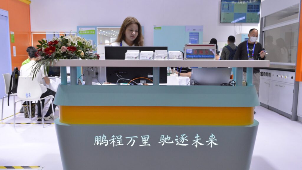 Pengchi Intelligent reception desk with flowers at the CHCC2025 booth in Wuhan International Expo Center, a staff member welcoming hospital construction professionals with product-category signage on the wall behind