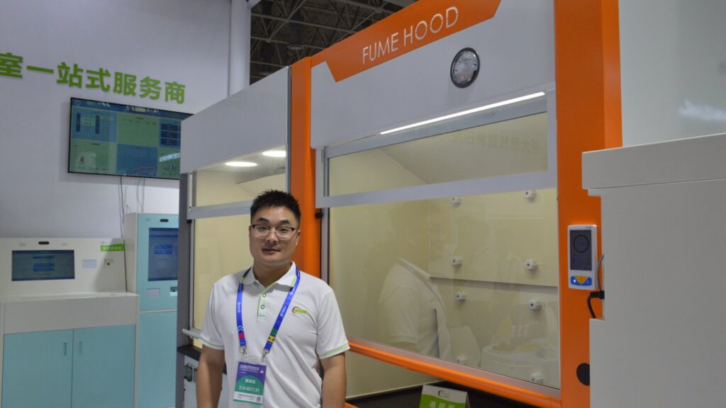 Pengchi engineer wearing an exhibitor badge standing beside an orange and white walk-in laboratory fume hood with glass sash, differential pressure gauge and exhaust controller at the CHCC2025 booth
