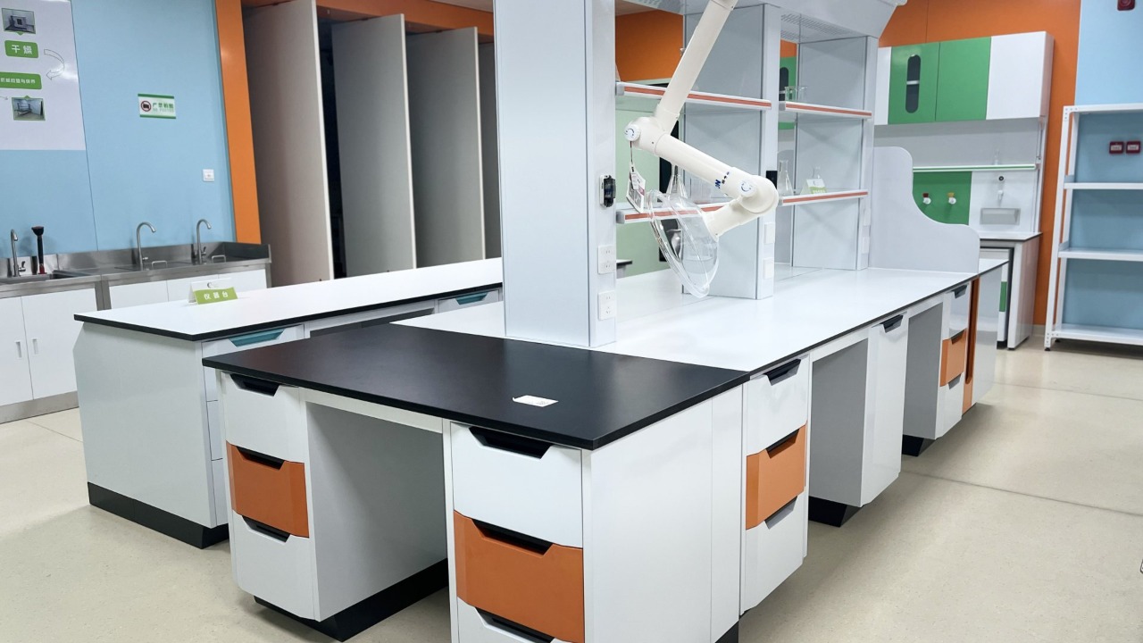 Un laboratoire moderne, propre et bien éclairé présente une longue rangée de paillasses centrales aux plateaux lisses, blancs et noirs mats, résistants aux produits chimiques, des armoires de rangement blanches rehaussées de façades de tiroirs orange vif, de poignées encastrées noires et de plinthes noires. Un bras articulé blanc d'extraction des fumées, équipé d'une hotte de captage transparente, est fixé à une colonne de service centrale, avec des étagères supérieures en verre accueillant de la verrerie de laboratoire plus loin le long de la rangée de paillasses. Des murs bleu pâle et orange encadrent des blocs éviers en acier inoxydable à gauche, des panneaux techniques arrière verts et blancs, un revêtement de sol beige sans joint et des rayonnages ouverts en métal blanc vides à droite, avec de petits panneaux de sécurité verts affichés dans cet espace épuré et fraîchement achevé.