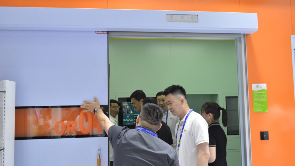 Visitors examining Pengchi's automatic hermetic operating room door marked OR 01 at CHCC2025, with a during-the-operation status sign and a surgical timer touchscreen panel inside the mocked-up operating room