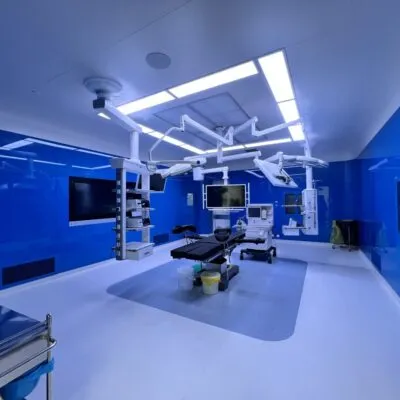 Une salle d'opération chirurgicale stérile, immaculée et inutilisée, présente des murs sans joint bleu profond très brillants et un sol lisse sans joint blanc éclatant, centrée sur une table d'opération réglable rembourrée noire posée sur un tapis de sol arrondi gris mat. Des bras articulés blancs montés au plafond suspendent des éclairages chirurgicaux, des moniteurs d'affichage et des étagères d'équipement, tandis que des panneaux LED linéaires encastrés lumineux diffusent un éclairage froid et uniforme dans tout l'espace clinique. Des écrans muraux, des armoires de rangement avec façade en verre bleu sur le côté droit, des chariots médicaux à roulettes et de petites poubelles à couvercle à côté de la table complètent ce bloc opératoire vide, ultra-propre, moderne et entièrement équipé.